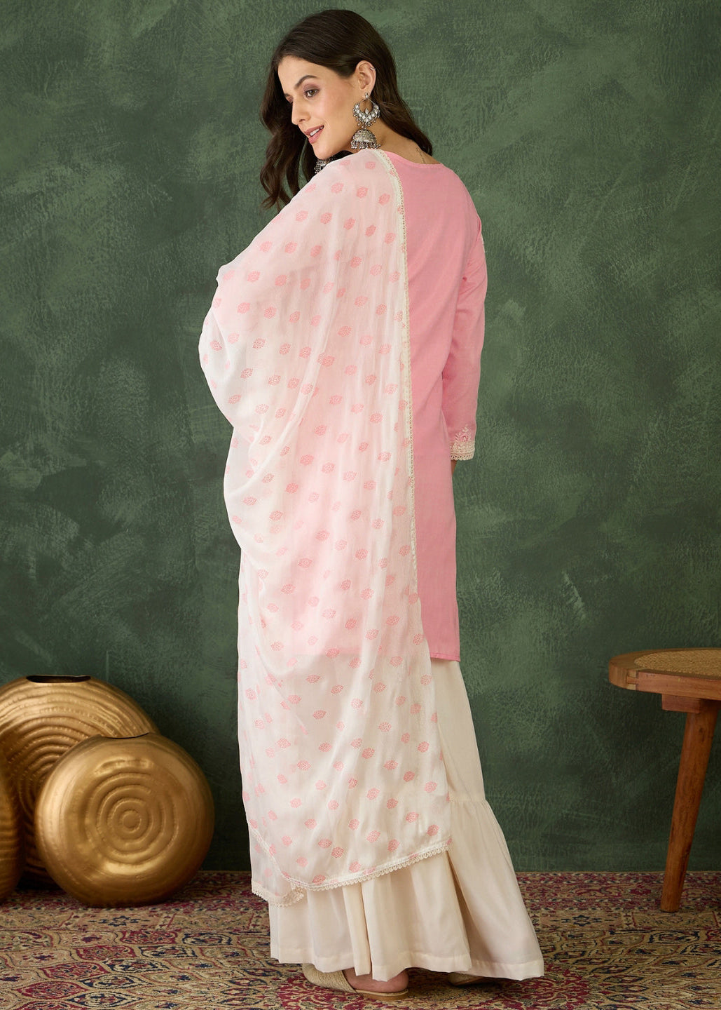 Woman wearing a pink and white saree against a green textured wall.