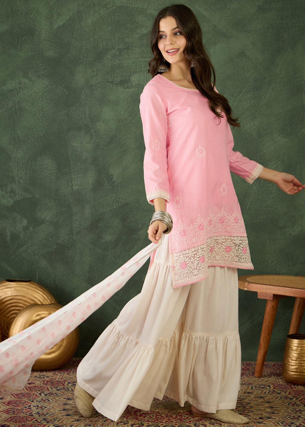 Woman wearing a pink embroidered top and white skirt against a green wall.