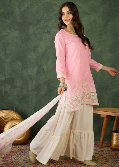 Woman wearing a pink embroidered top and white skirt against a green wall.