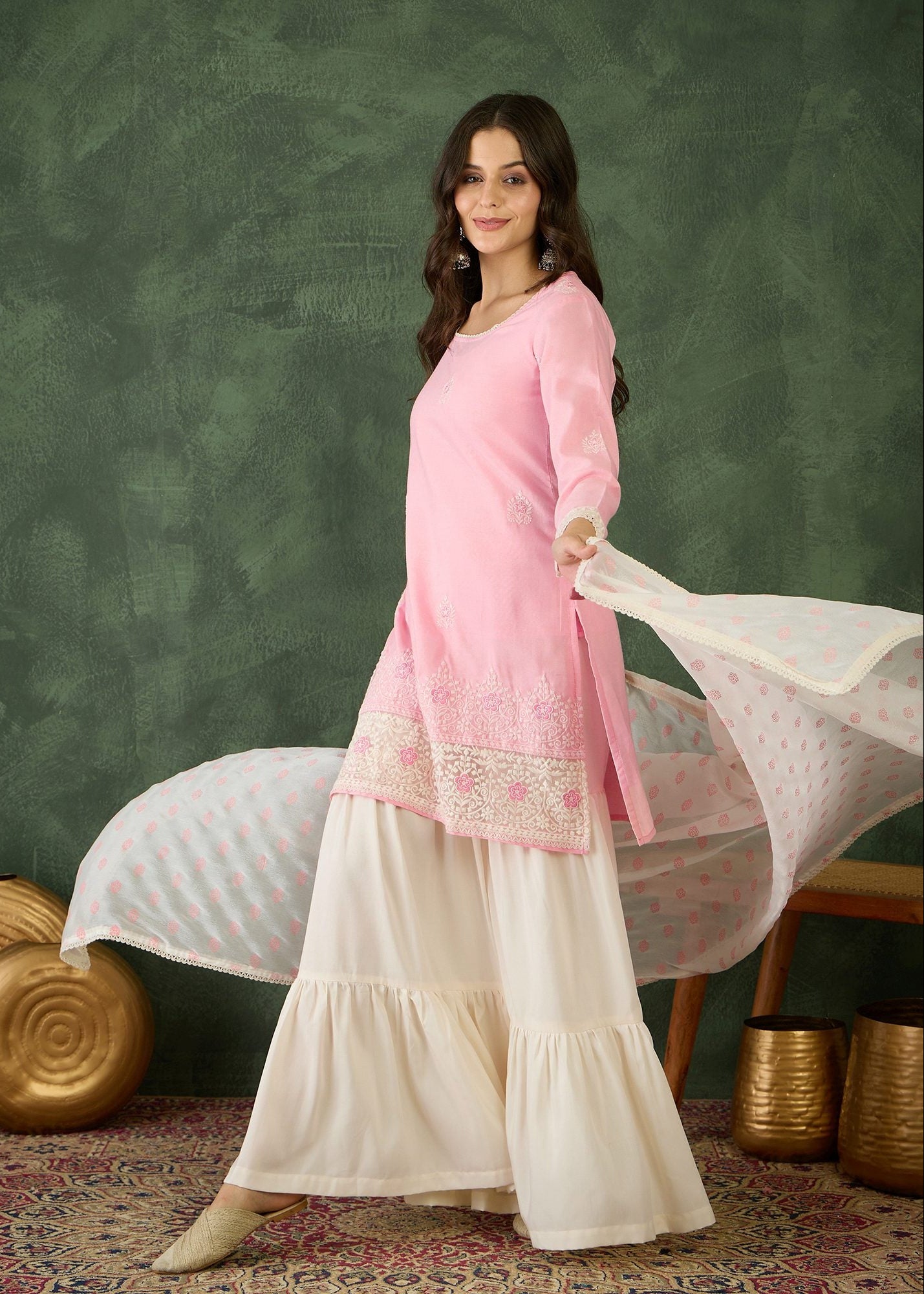Woman in a pink top and white pants holding a scarf against a green textured wall.