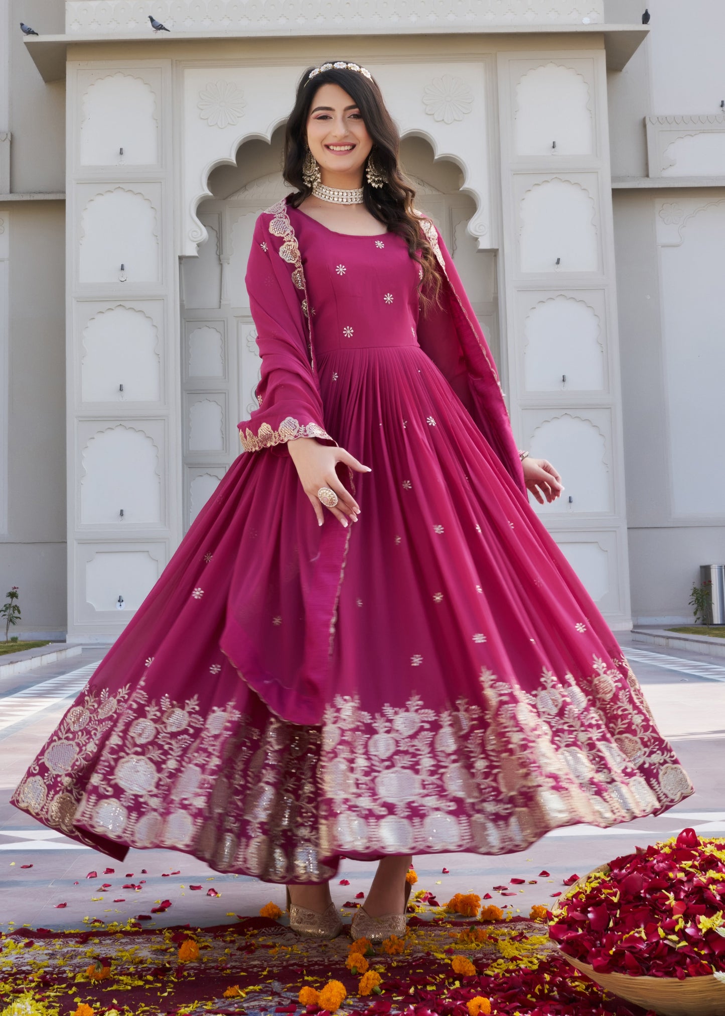 Woman in a pink traditional outfit standing in front of a decorative archway with flowers on the ground.