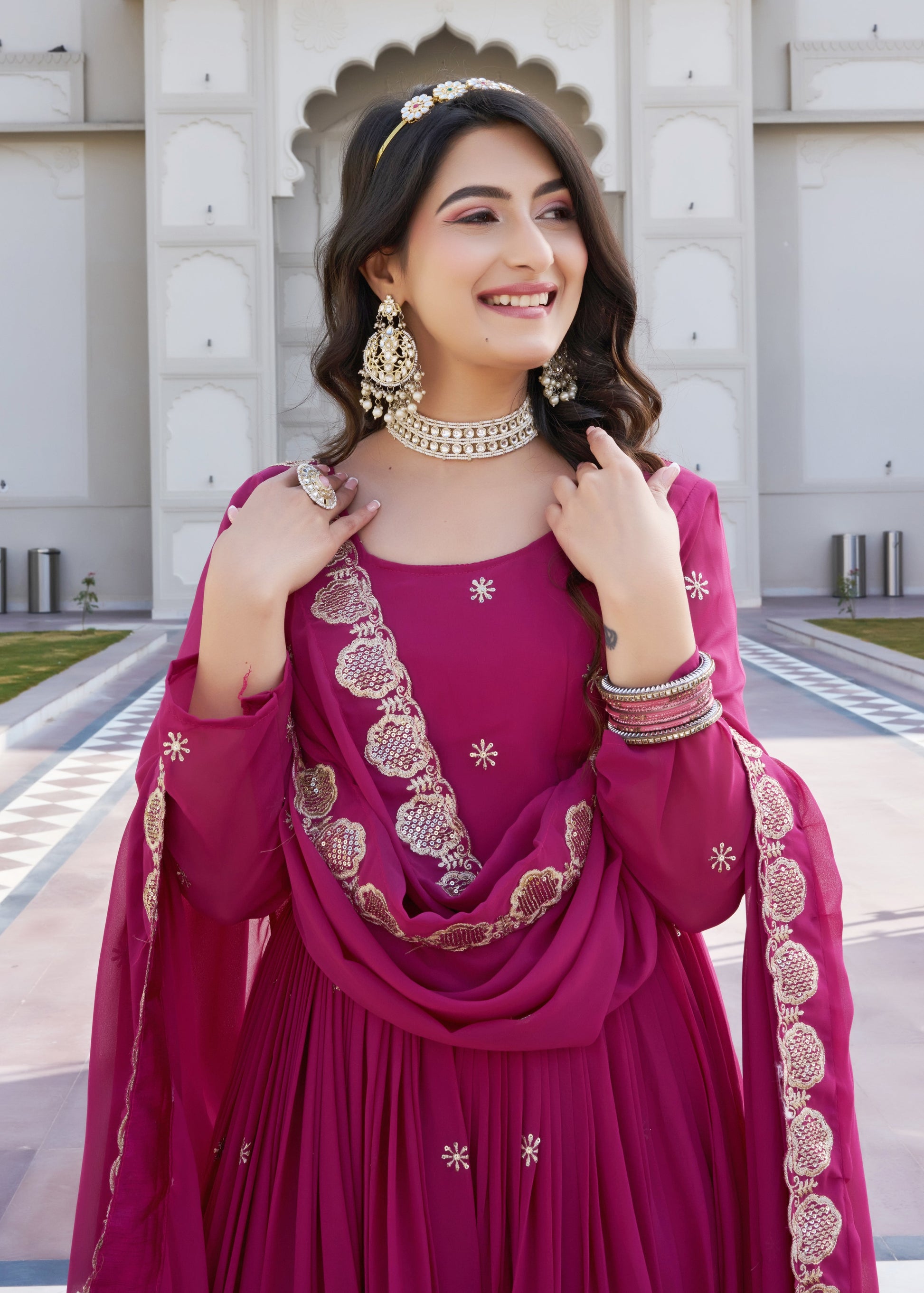 Woman in a traditional pink outfit with gold embroidery standing in front of a decorative wall.