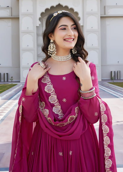 Woman in a traditional pink outfit with gold embroidery standing in front of a decorative wall.